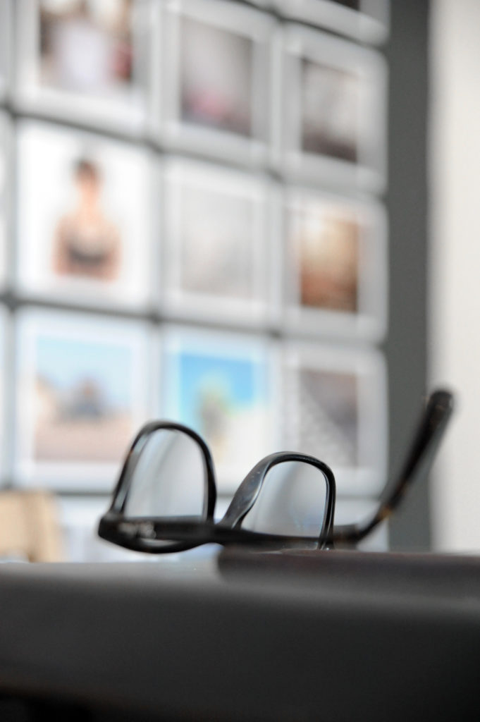 a pair of glasses on the edge of a table with blurred out grid of photographs hung on the wall behind.